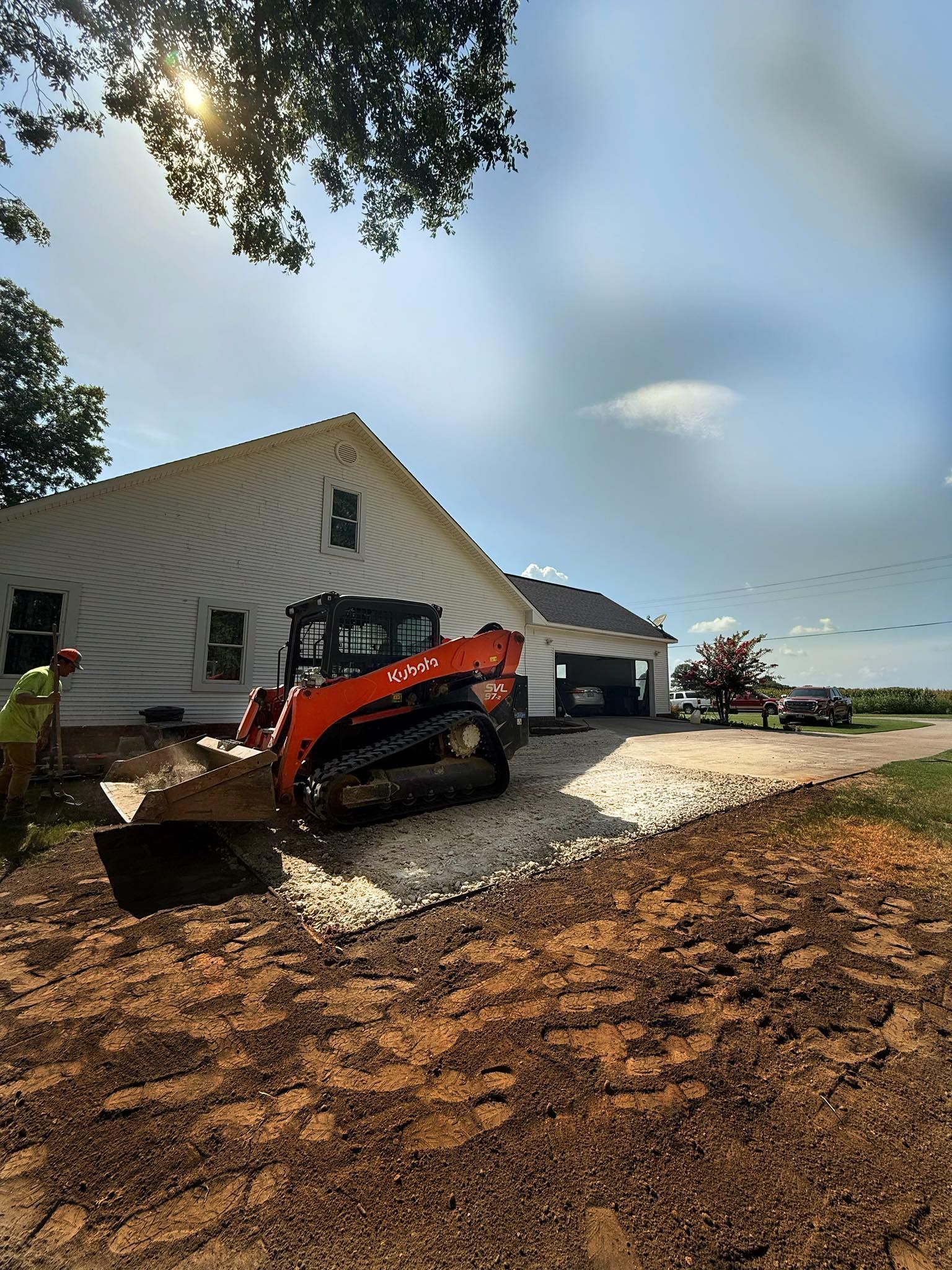An orange skid steer with tracks moves white gravel in front of a white house on a sunny day.