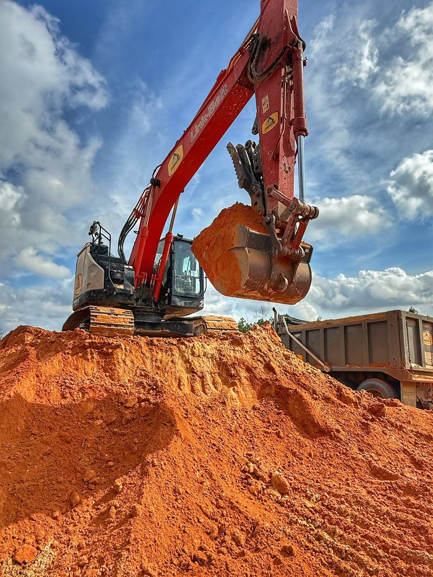 A red excavator sits atop a pile of orange dirt, lifting a bucket full of soil near a dump truck against a cloudy sky.