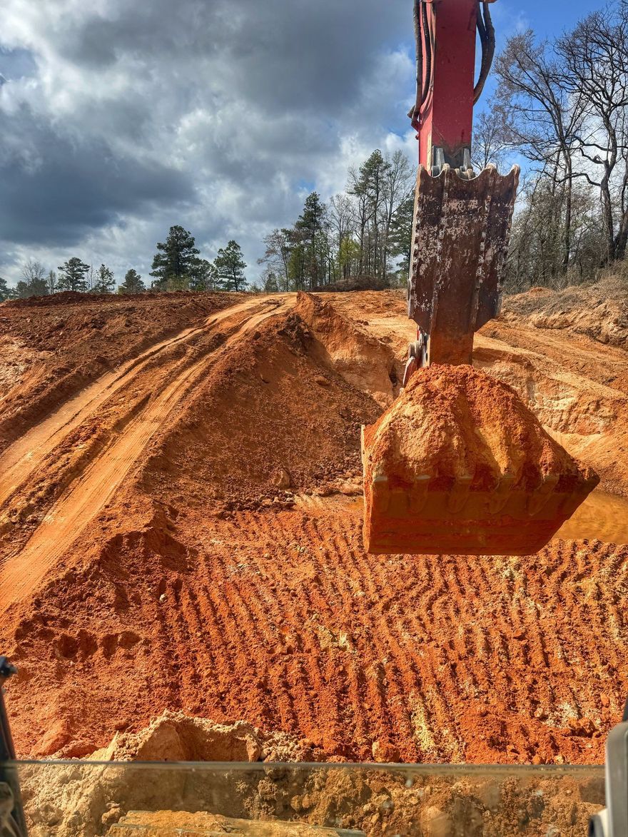 An excavator bucket filled with red clay dirt suspended over an active construction site with trees and a cloudy sky.