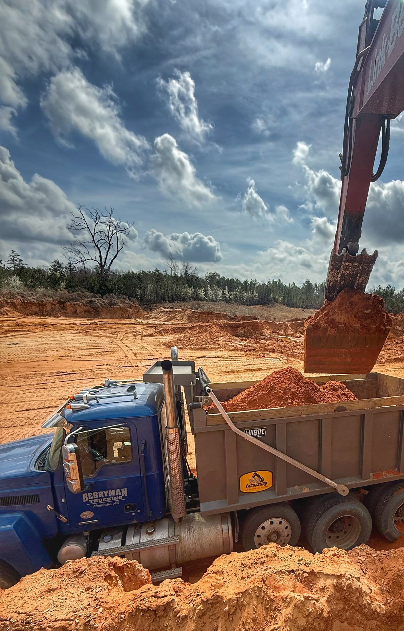 An excavator filling a blue dump truck with dirt at a construction site under a cloudy sky.
