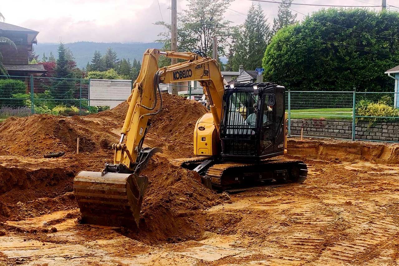 A yellow excavator digs into a large pile of dirt at an active construction site with trees in the background.