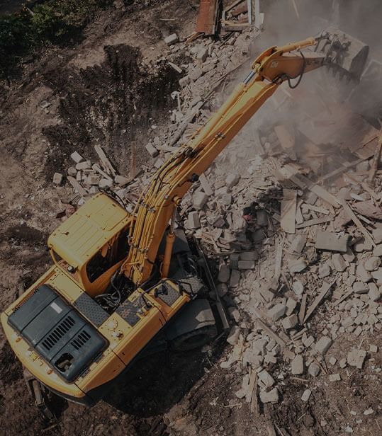 An aerial view of a yellow excavator demolishing a pile of concrete rubble at a construction site.