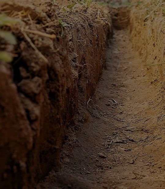 A narrow, straight trench cut into dry, brown soil, with rough dirt walls showing layers of earth and roots.