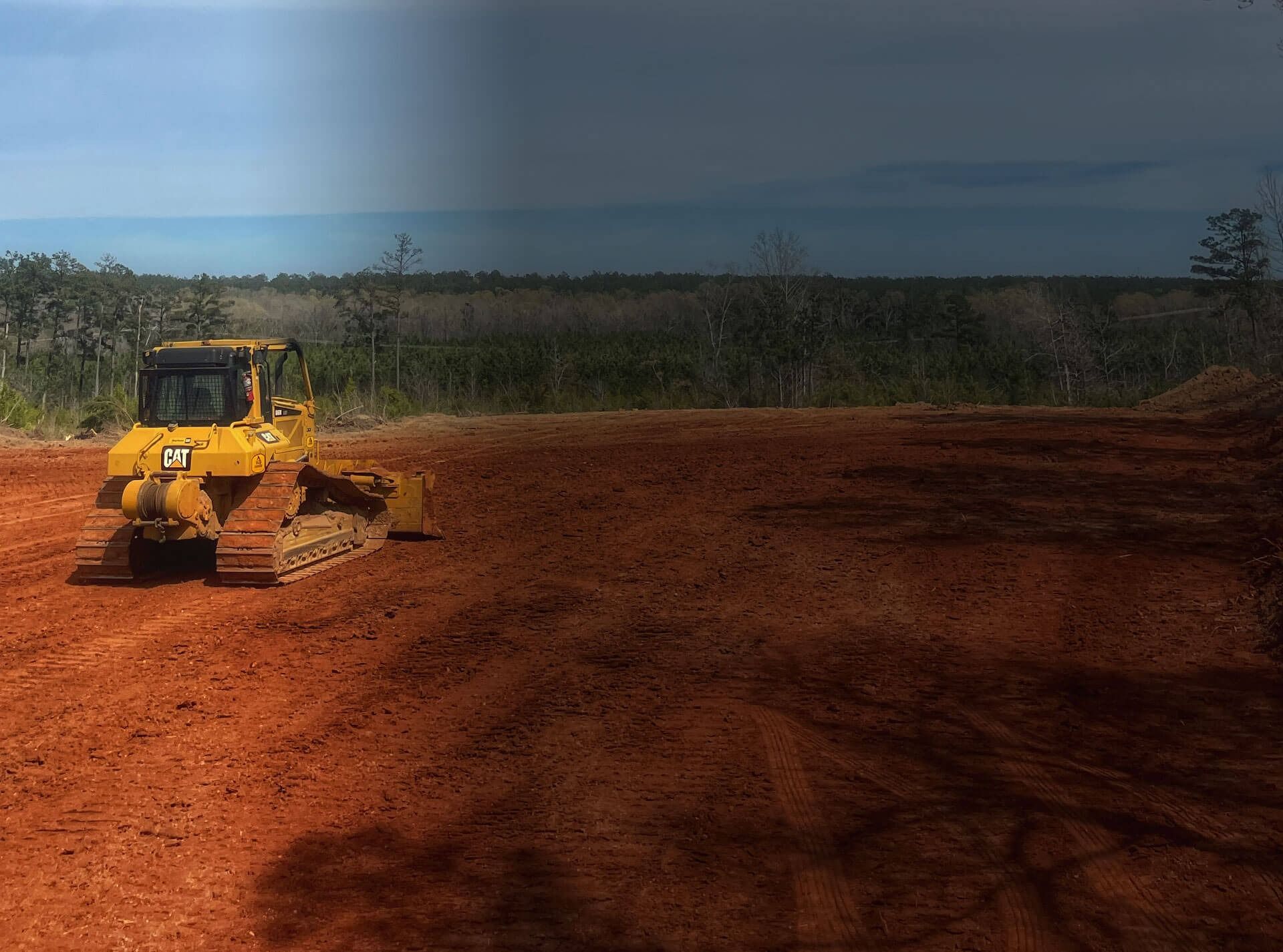 A yellow bulldozer parked on a flat, reddish-brown dirt clearing with a forested landscape in the background.