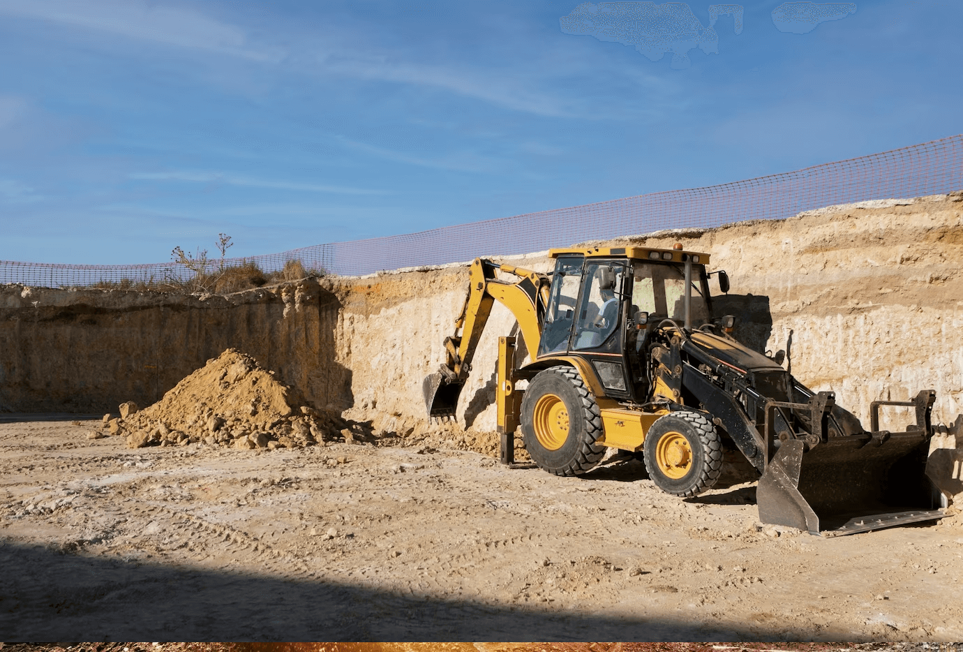 A yellow backhoe loader sits in an open dirt quarry under a blue sky, next to a pile of excavated soil.