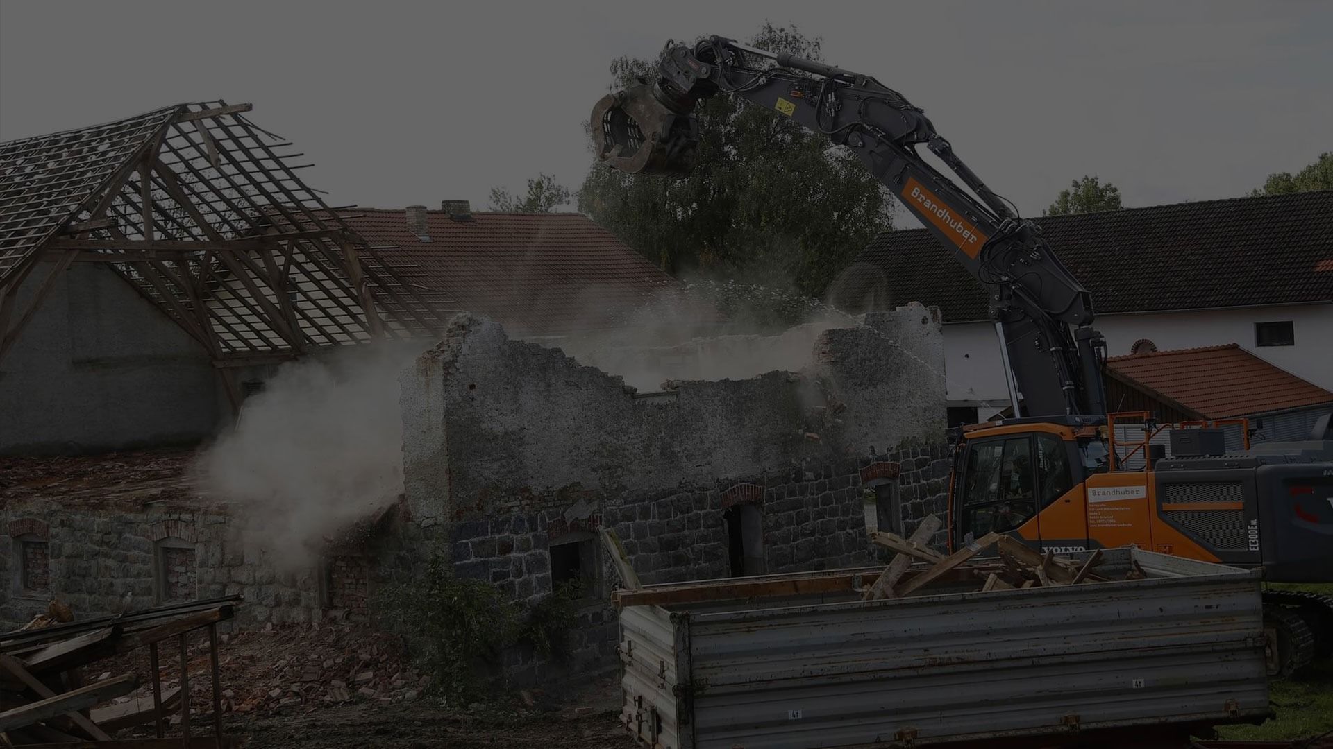An excavator demolishing an old stone structure, creating a cloud of dust amid debris and a wooden frame building.