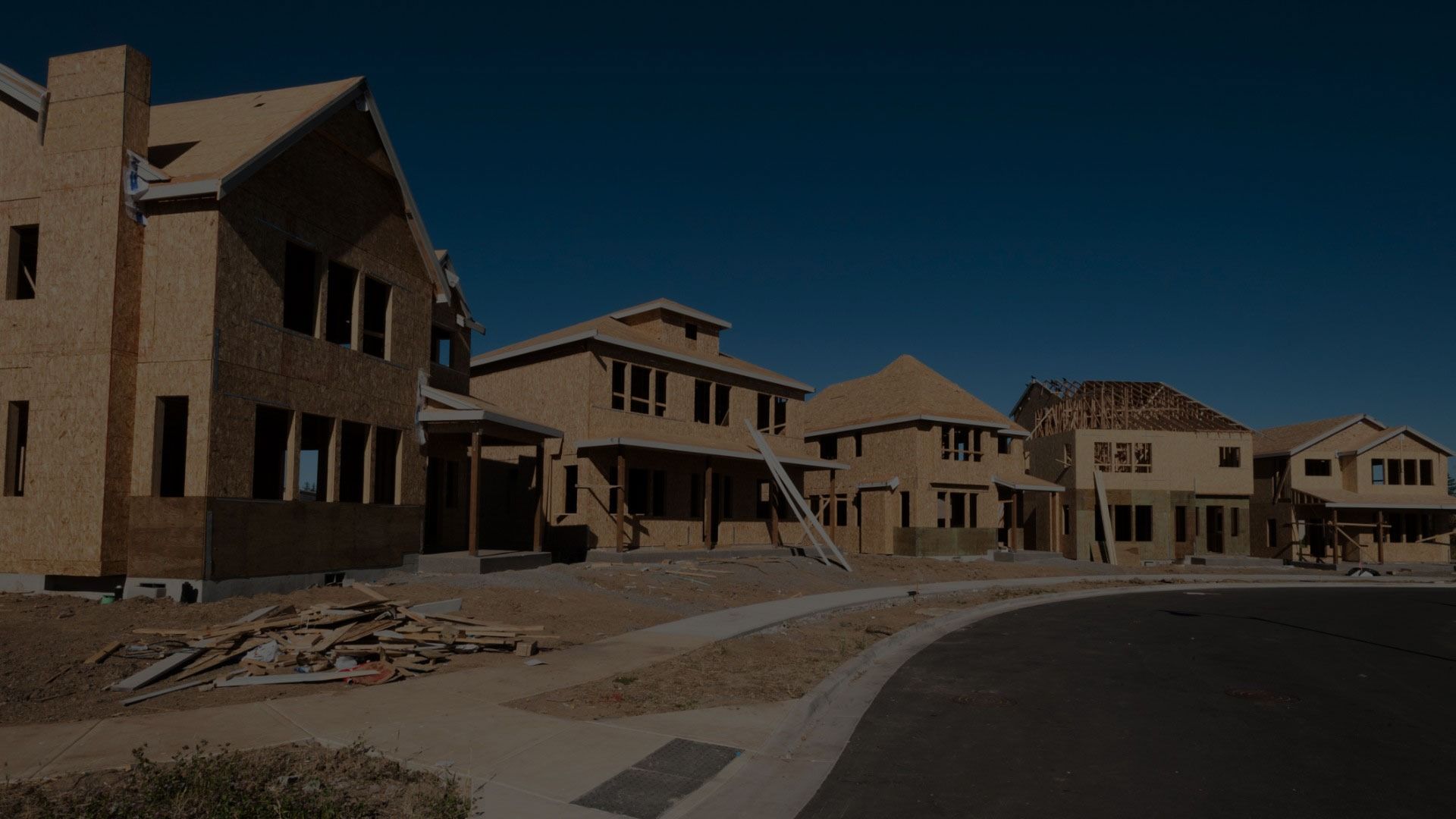 A row of unfinished wooden houses under construction in a suburban neighborhood under a clear blue sky.