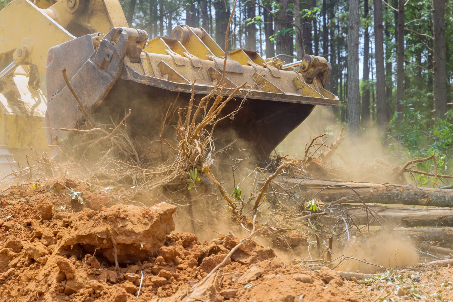 A yellow excavator bucket clearing red dirt and debris in a forest.