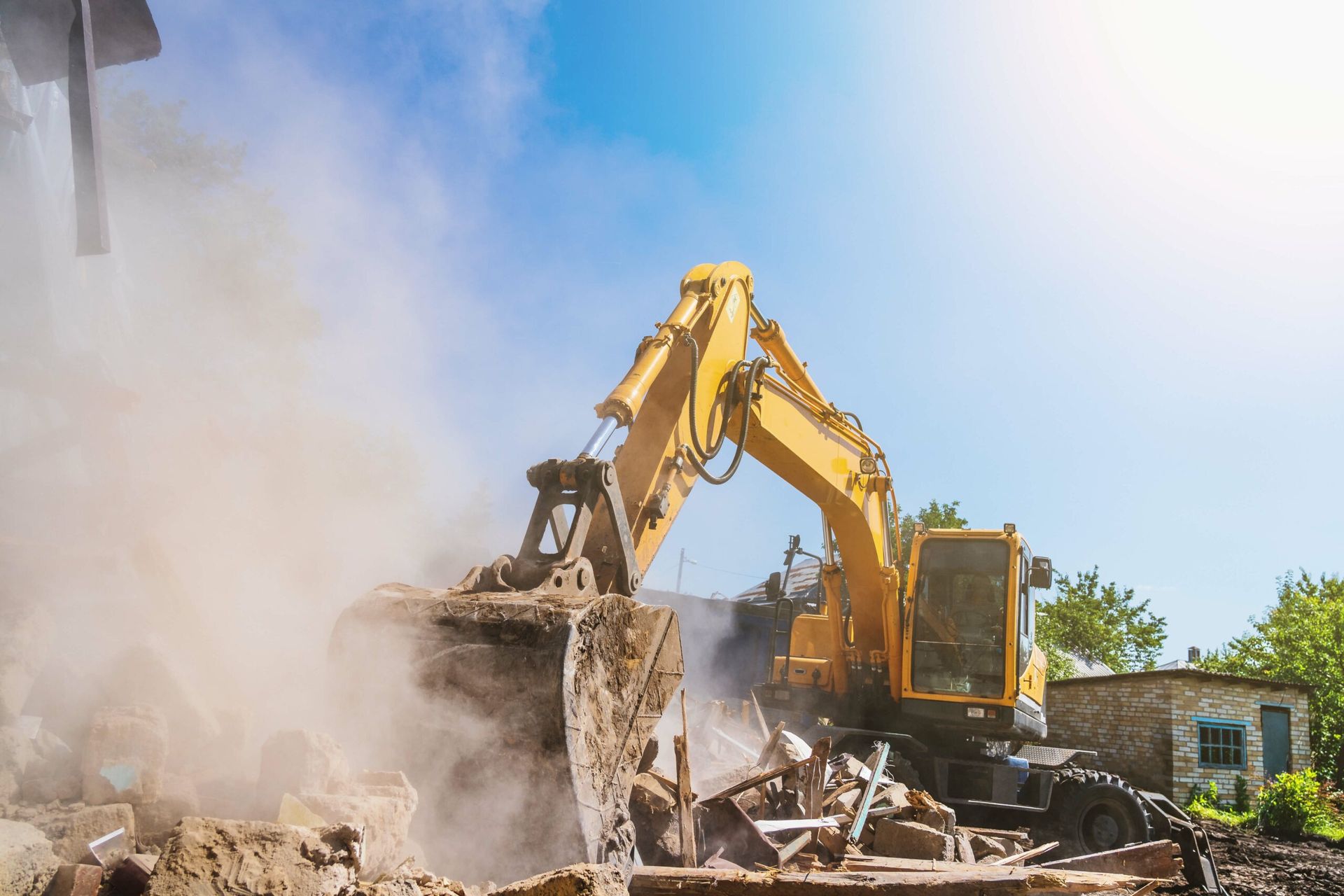 A yellow excavator demolishes a structure, kicking up a large cloud of dust under a sunny blue sky.