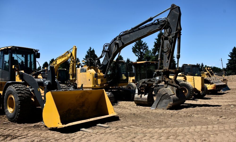 A row of yellow and grey construction vehicles, including excavators and a loader, parked on a dirt site under a blue sky.