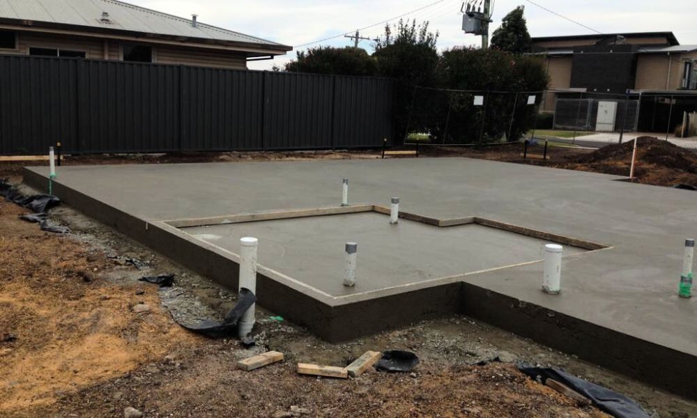 Newly poured grey concrete slab foundation for a building with white plumbing pipes protruding from the surface.