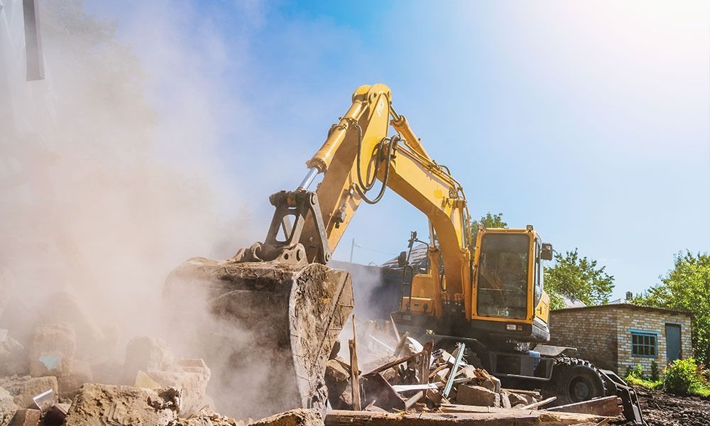 A yellow excavator tears through a building, kicking up a large cloud of dust under a clear, bright blue sky.