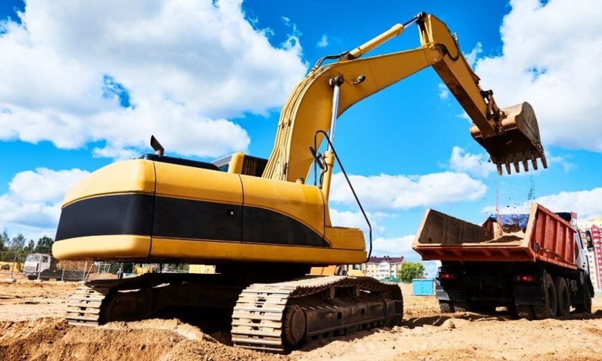 Yellow excavator loading dirt into a dump truck at a construction site against a blue sky with clouds.