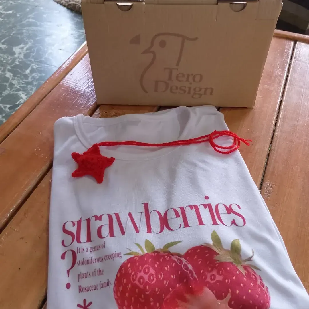 Camiseta blanca con gráfico de fresa, adorno de estrella roja y una caja etiquetada