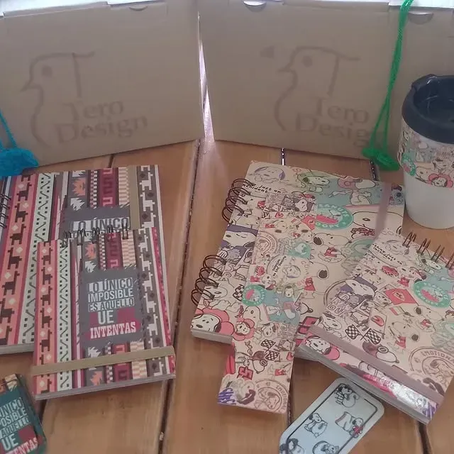 Cuadernos y taza de viaje sobre una mesa de madera, cajas en el fondo, todo con el logo del pájaro.