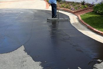 A person is spreading asphalt on a driveway with a broom.