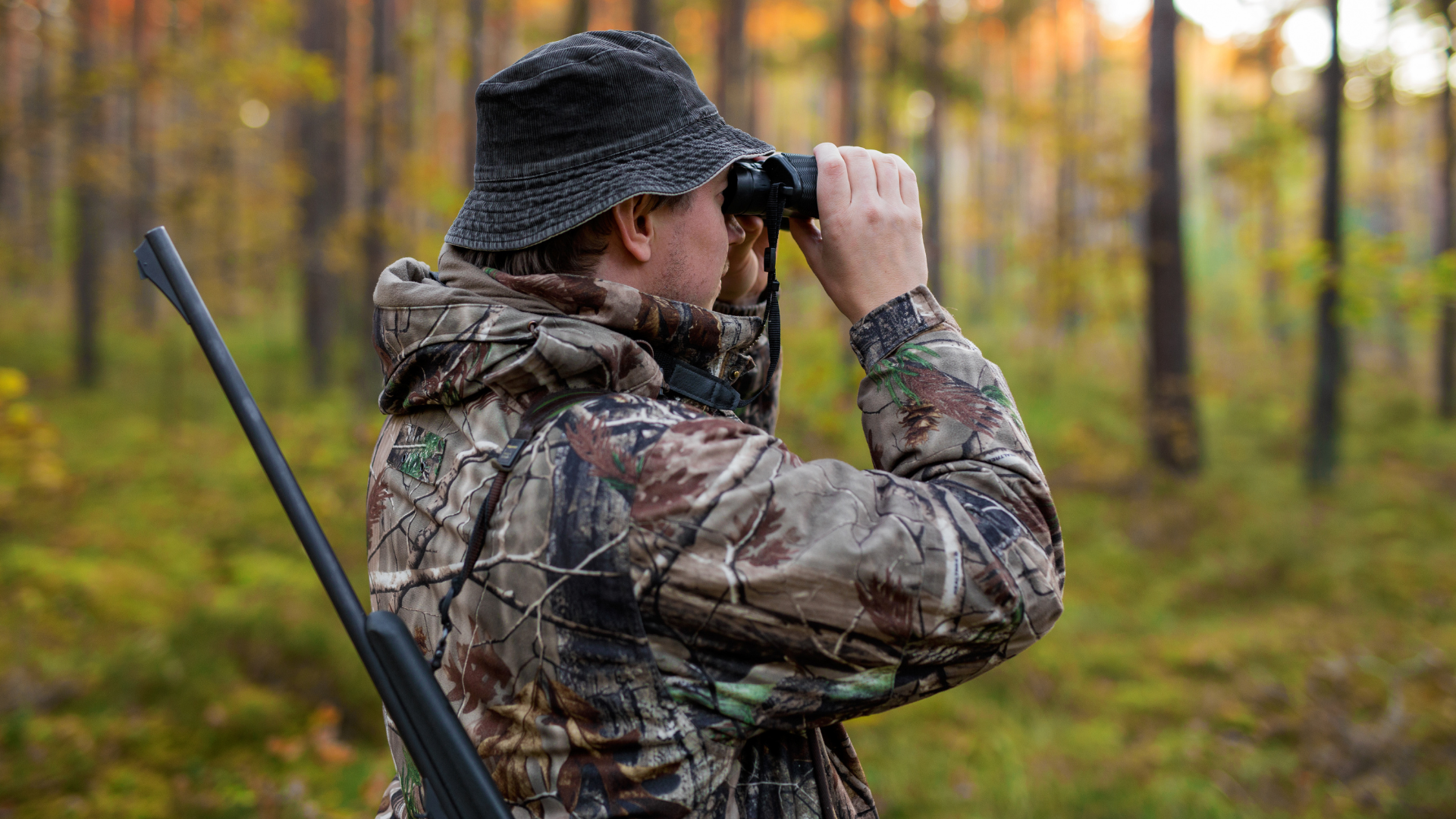Hunter in camouflage gear looking through binoculars in a forest.