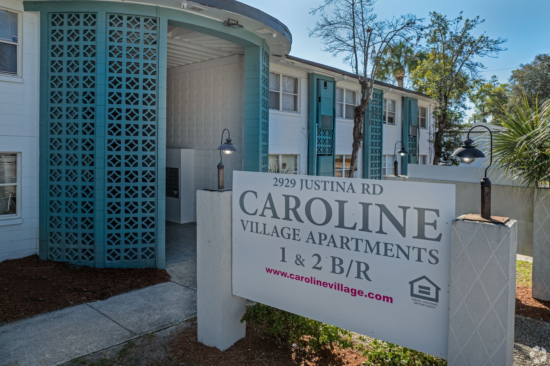Caroline Village Apartments entrance sign. Teal decorative screen, white buildings, blue sky.