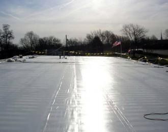 White roof with visible seams, reflecting sunlight. Trees and a flag in the background.