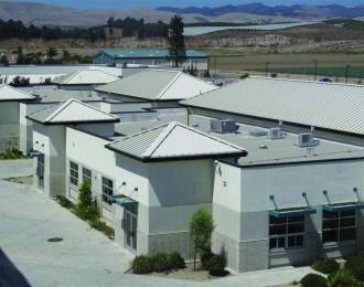 Buildings with light gray walls and white trim, metal roofs, and a distant field and hills.