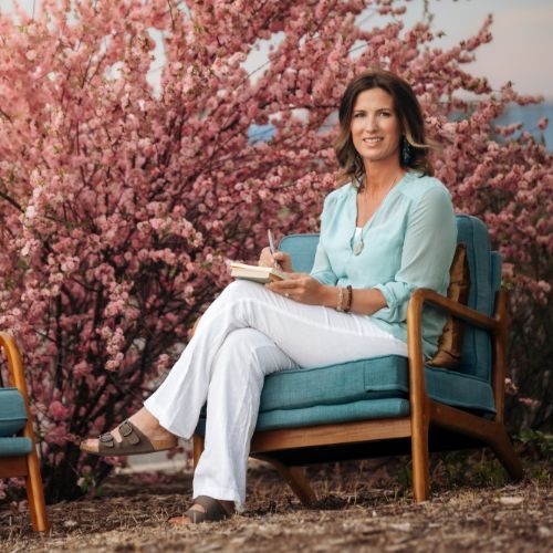 Woman writing in a notebook, sitting on a blue chair near pink flowering tree. She wears white pants and blue shirt.