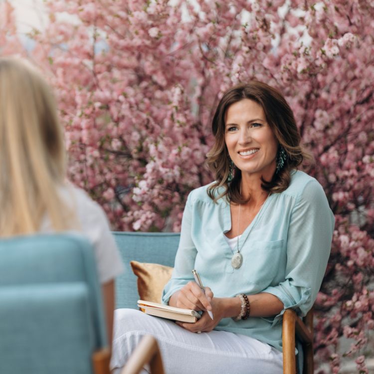 Woman in light blue top smiles, takes notes while talking to someone outdoors with pink flowers.