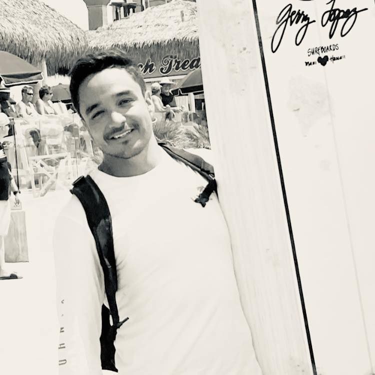 Man smiles while holding a surfboard on a beach with people and a thatch-roofed building in the background.