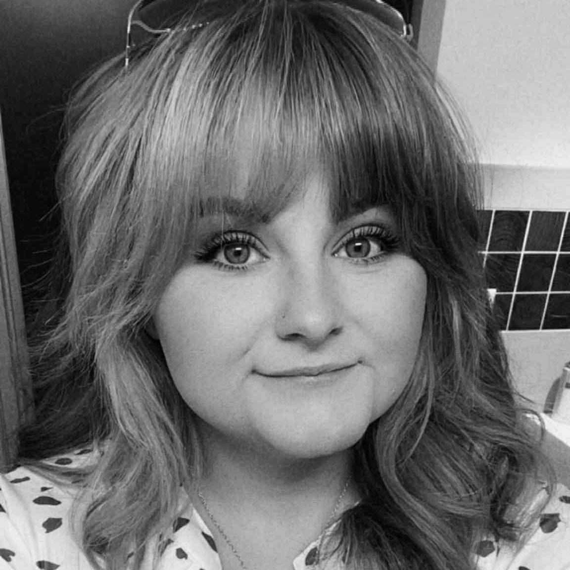 Woman with light hair, bangs, and smiling expression. Wearing a spotted shirt.