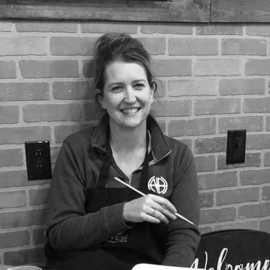 Woman smiling, holding paintbrush, in front of brick wall. Wearing apron and sweatshirt, indoors.