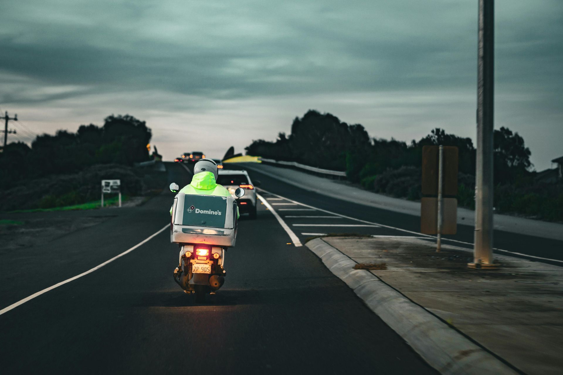 Un repartidor en motocicleta, con ropa reflectante, circula por una carretera al atardecer; un coche le sigue.