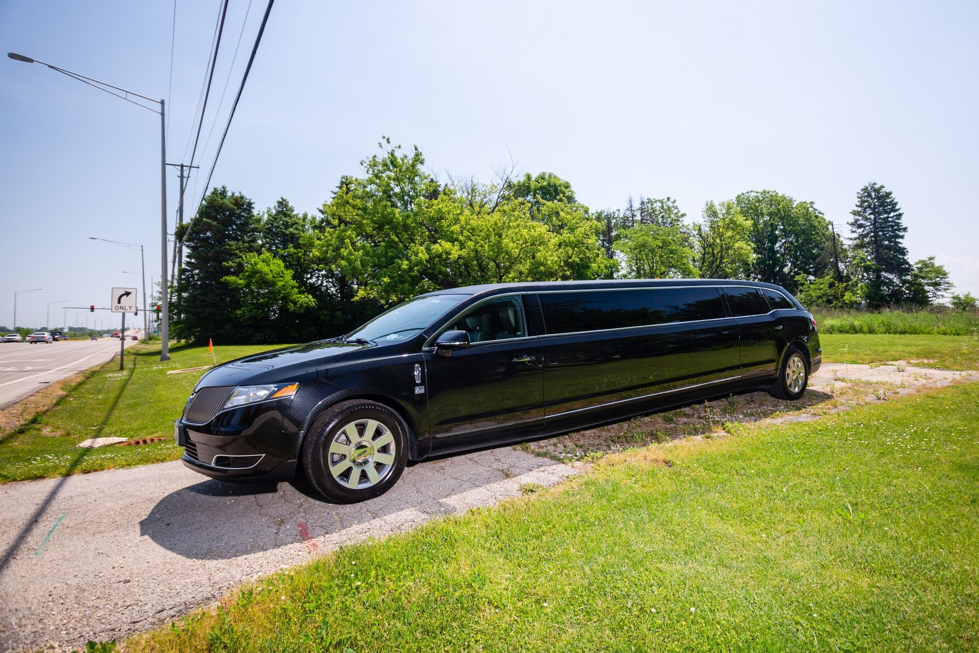 Black limousine parked on a paved area near a green grassy area with trees and power lines in the background.