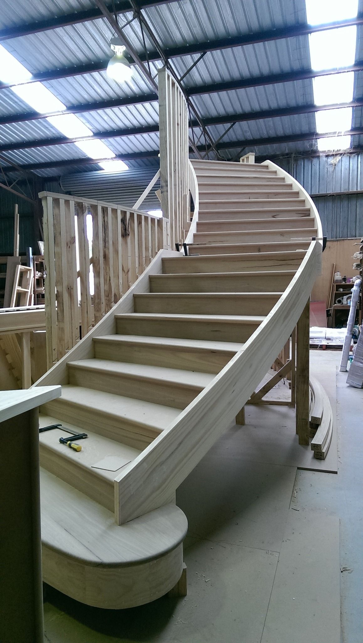 Wooden spiral staircase under construction inside a workshop with natural light.