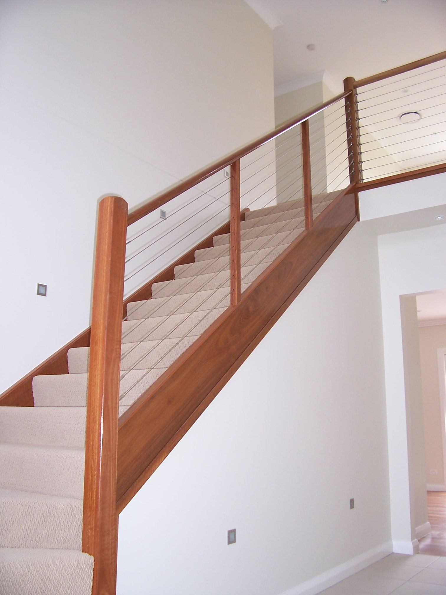 Wooden staircase with carpeted steps and metal cable railing in a white-walled home.