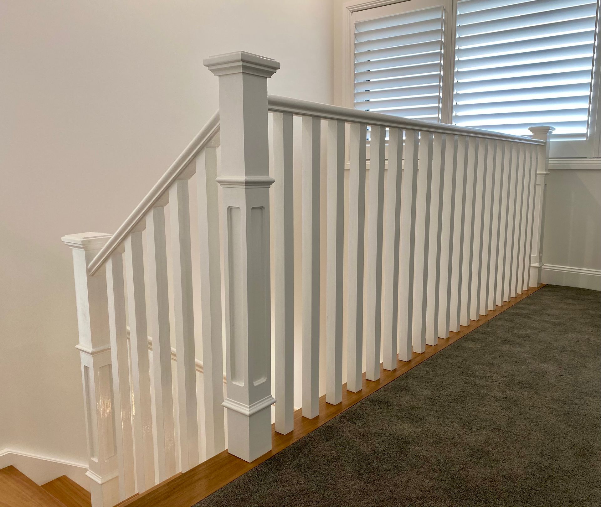 White stair railing with vertical spindles, next to carpeted area and window with shutters.