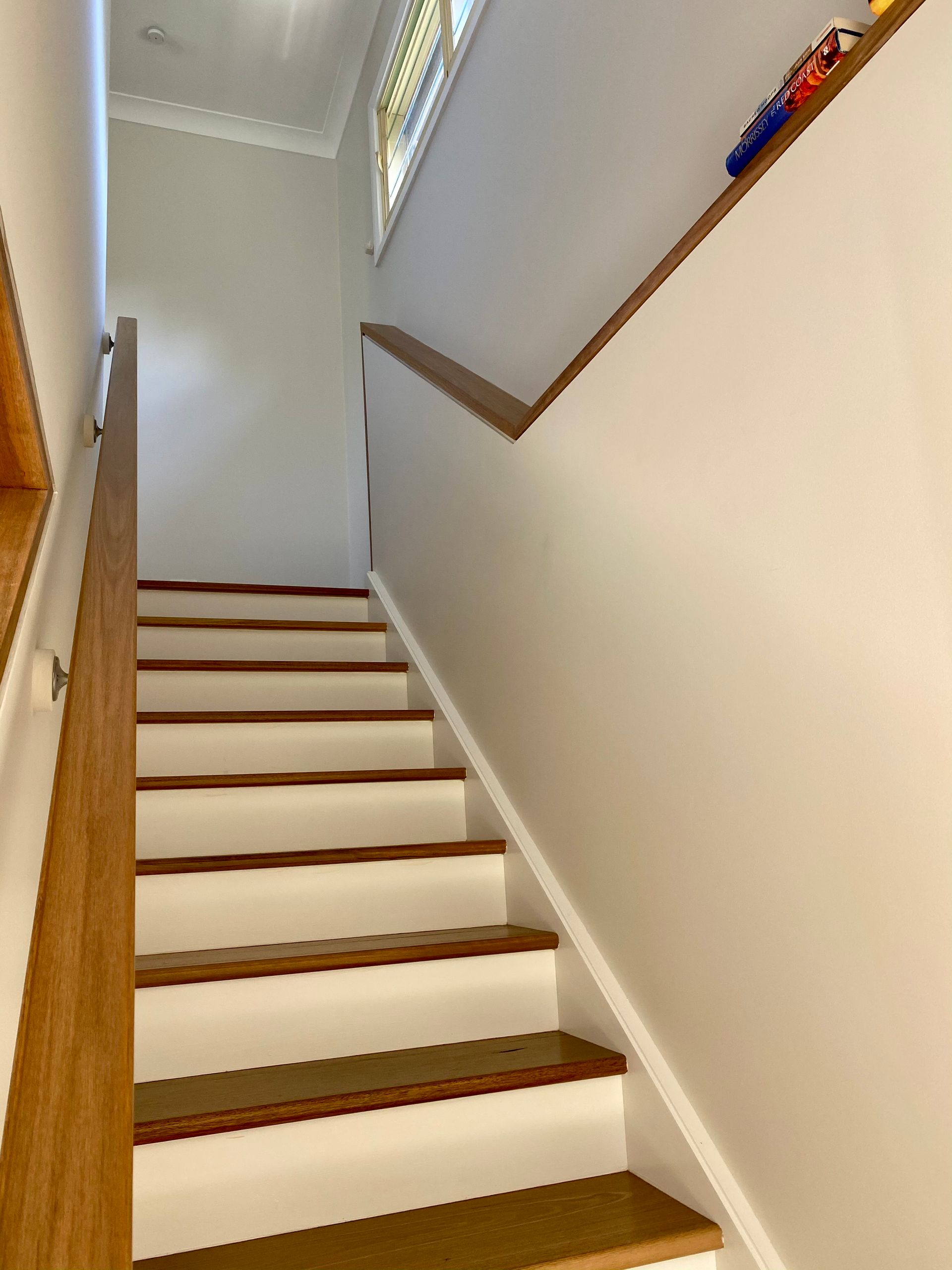 Staircase with white risers and wooden treads and handrails, leading upwards against a white wall.