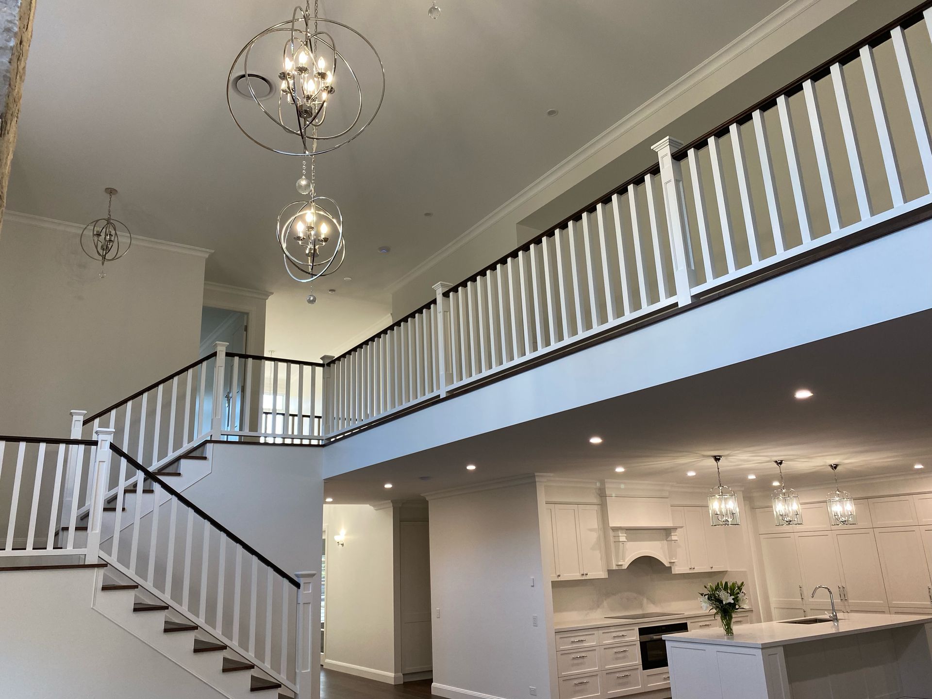 Two-story interior with a staircase and a kitchen visible. White walls, black railings, and chandeliers.