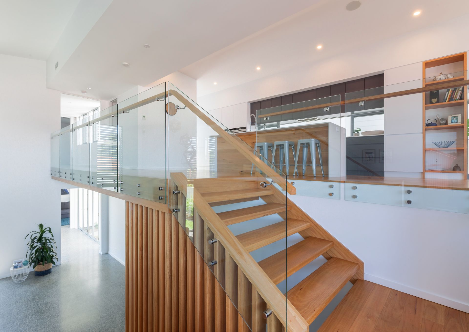 Wooden staircase with glass railing, leading to a mezzanine overlooking a bright living area.