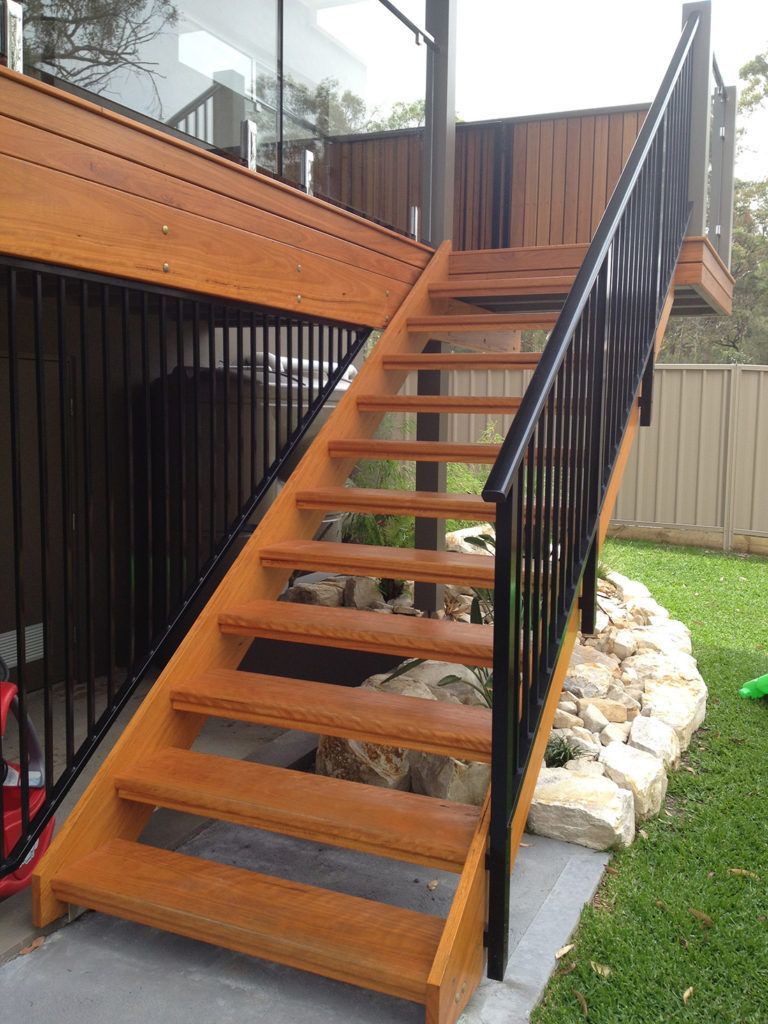 Wooden outdoor staircase with black railing leading to a deck, beside a lawn.