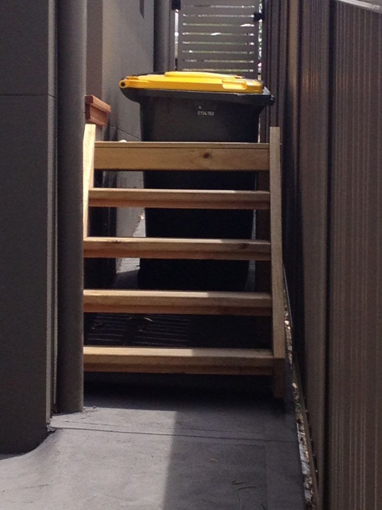 Wooden stairs leading up to a black and yellow trash bin in a narrow outdoor alley.
