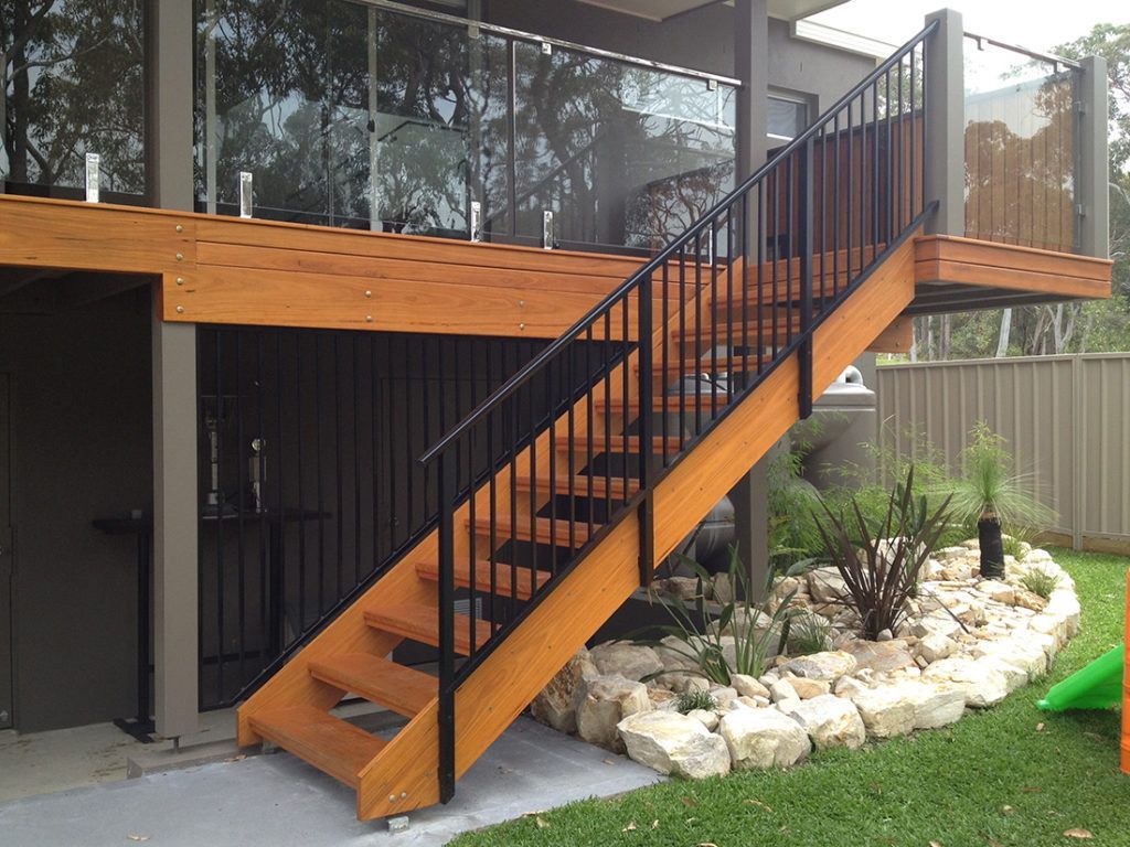 Wooden staircase with black railing leading to a deck. Outdoor setting with landscaping and a fence.