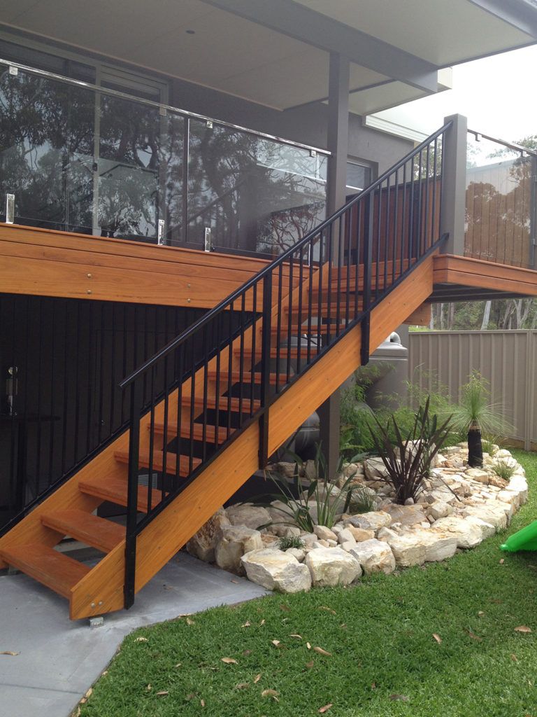 Wooden outdoor staircase with black metal railings, leading from a deck to a grassy yard with a rock garden.