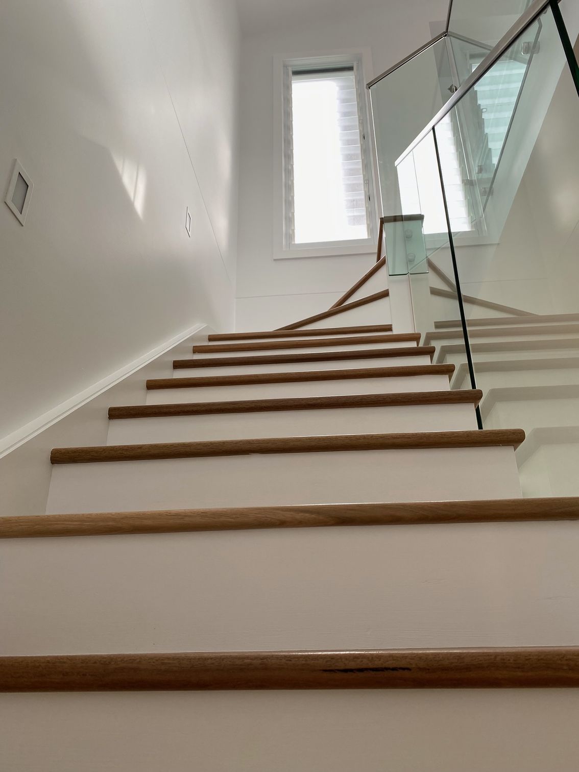Staircase with white risers, wooden treads and handrail, glass railing, and a window at the top.