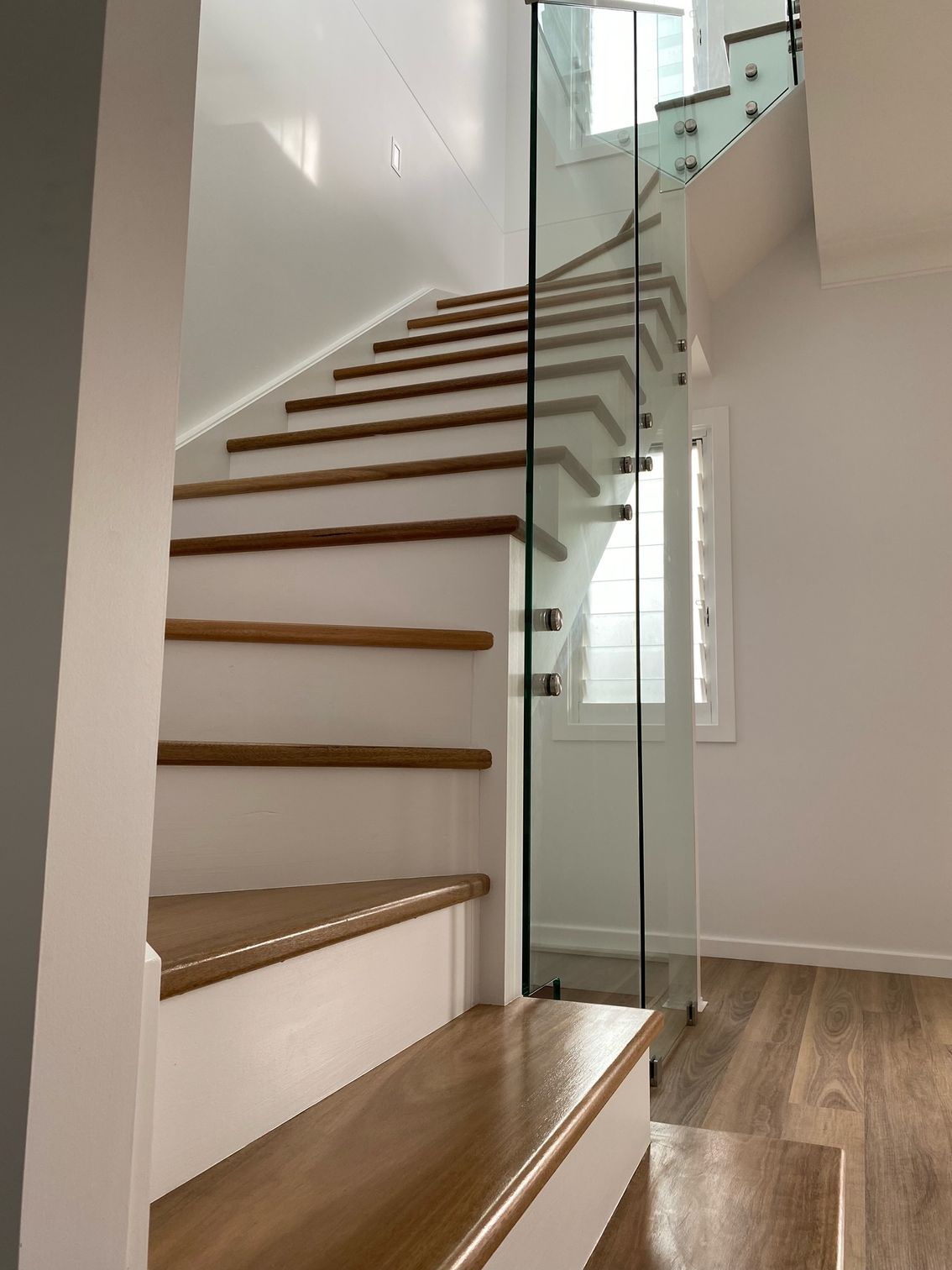 Wooden staircase with glass railing, ascending to the right, against a white wall.