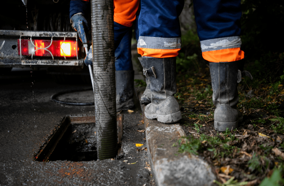 Workers cleaning a sewer, hose inserted into the open manhole, wearing reflective orange cuffs and boots.