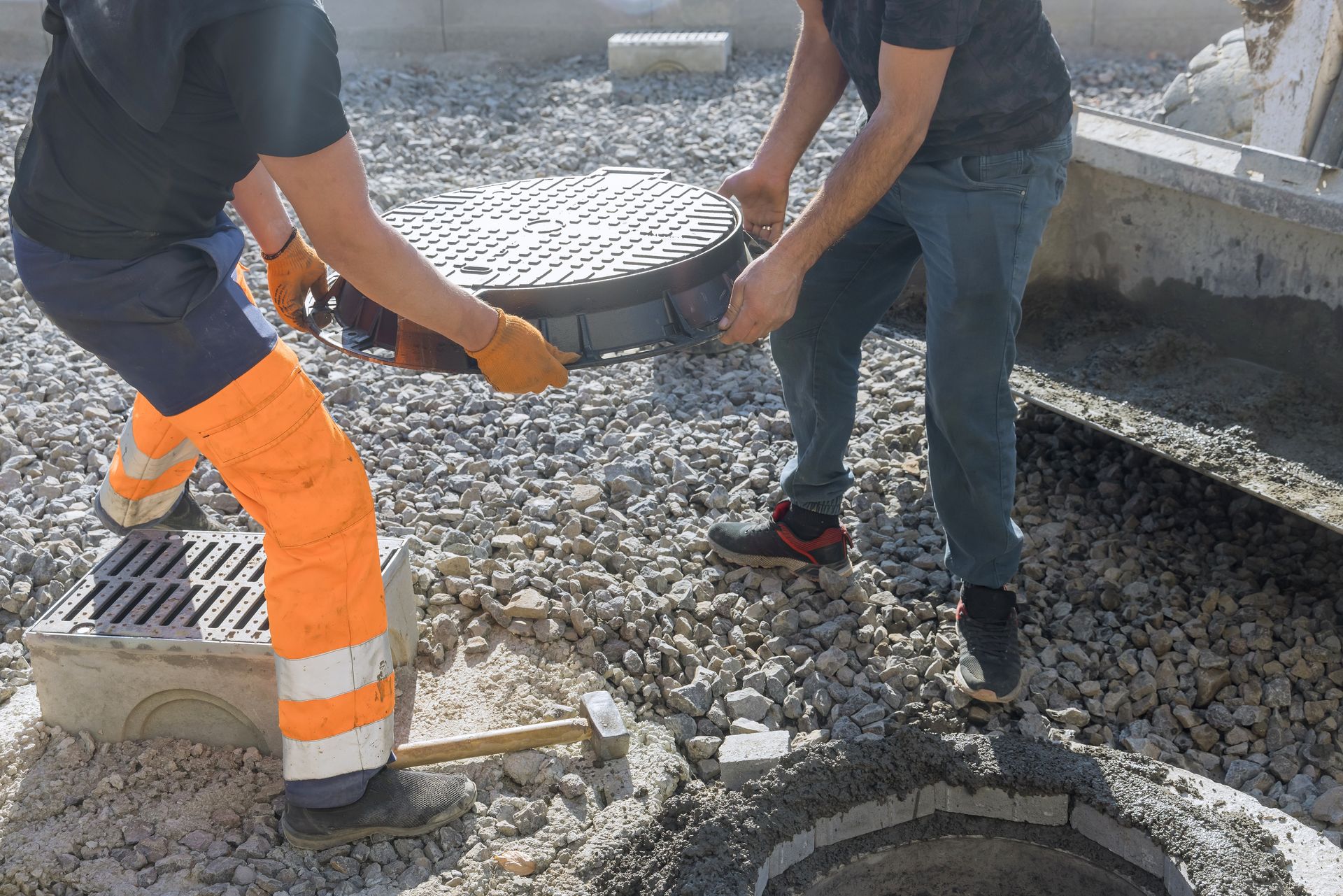 Two workers placing a manhole cover on a drain, on a gravel surface.