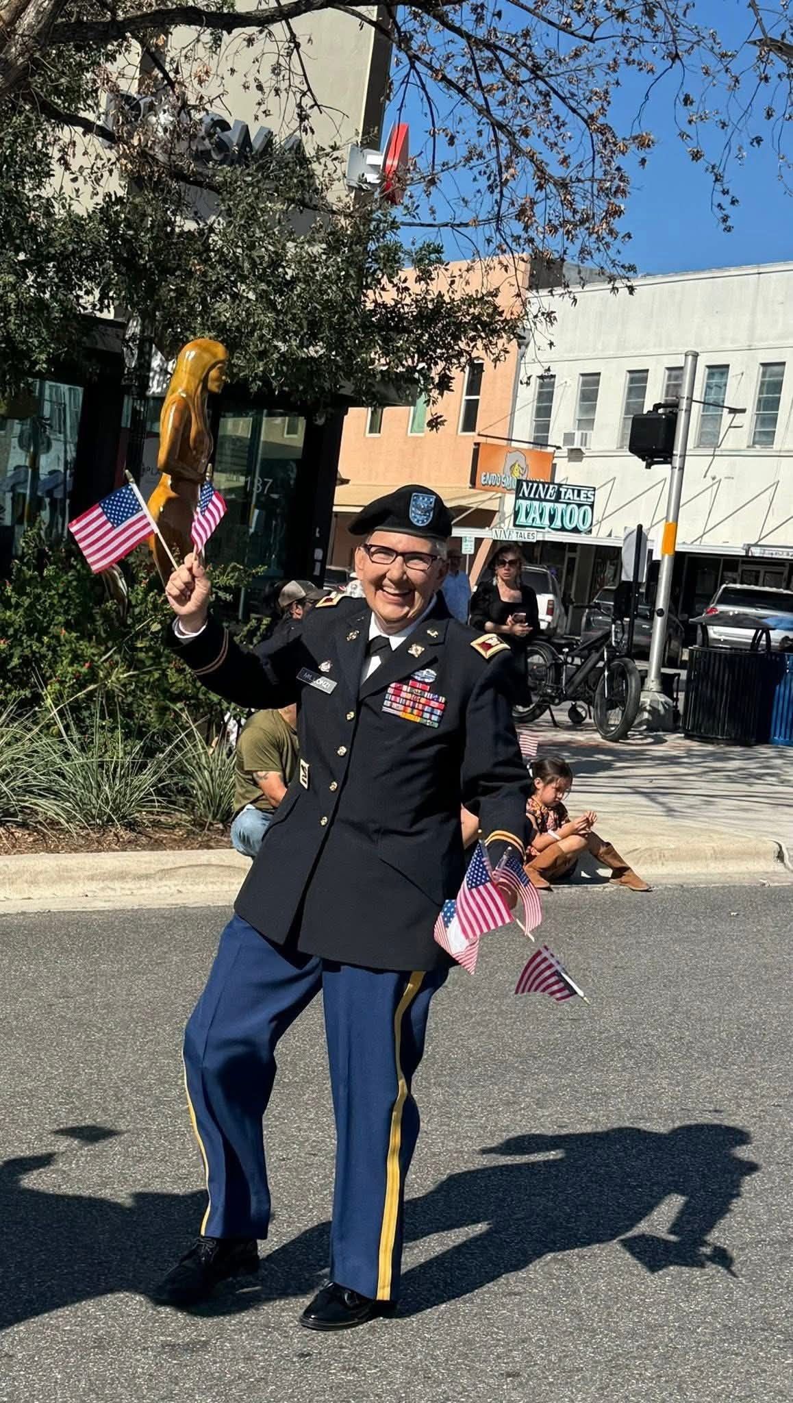 Man in military uniform waving American flags in a parade; buildings and trees in the background.