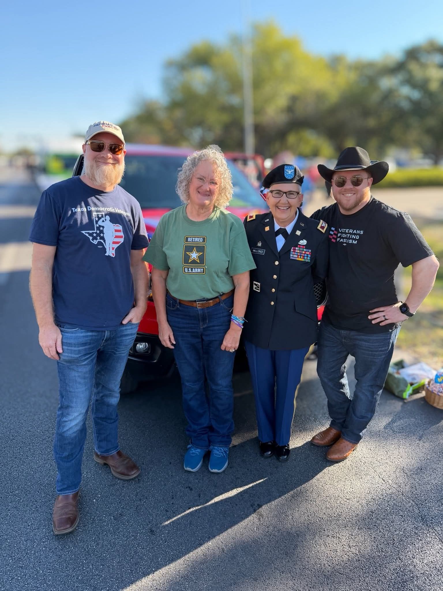 Four people pose outdoors, a military officer in uniform stands among them. They are in front of a red vehicle.