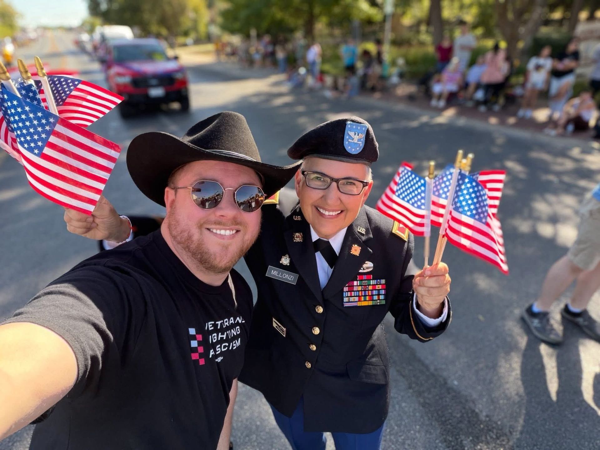 Two people smiling and waving American flags at a parade. One wears a cowboy hat, the other a military uniform.