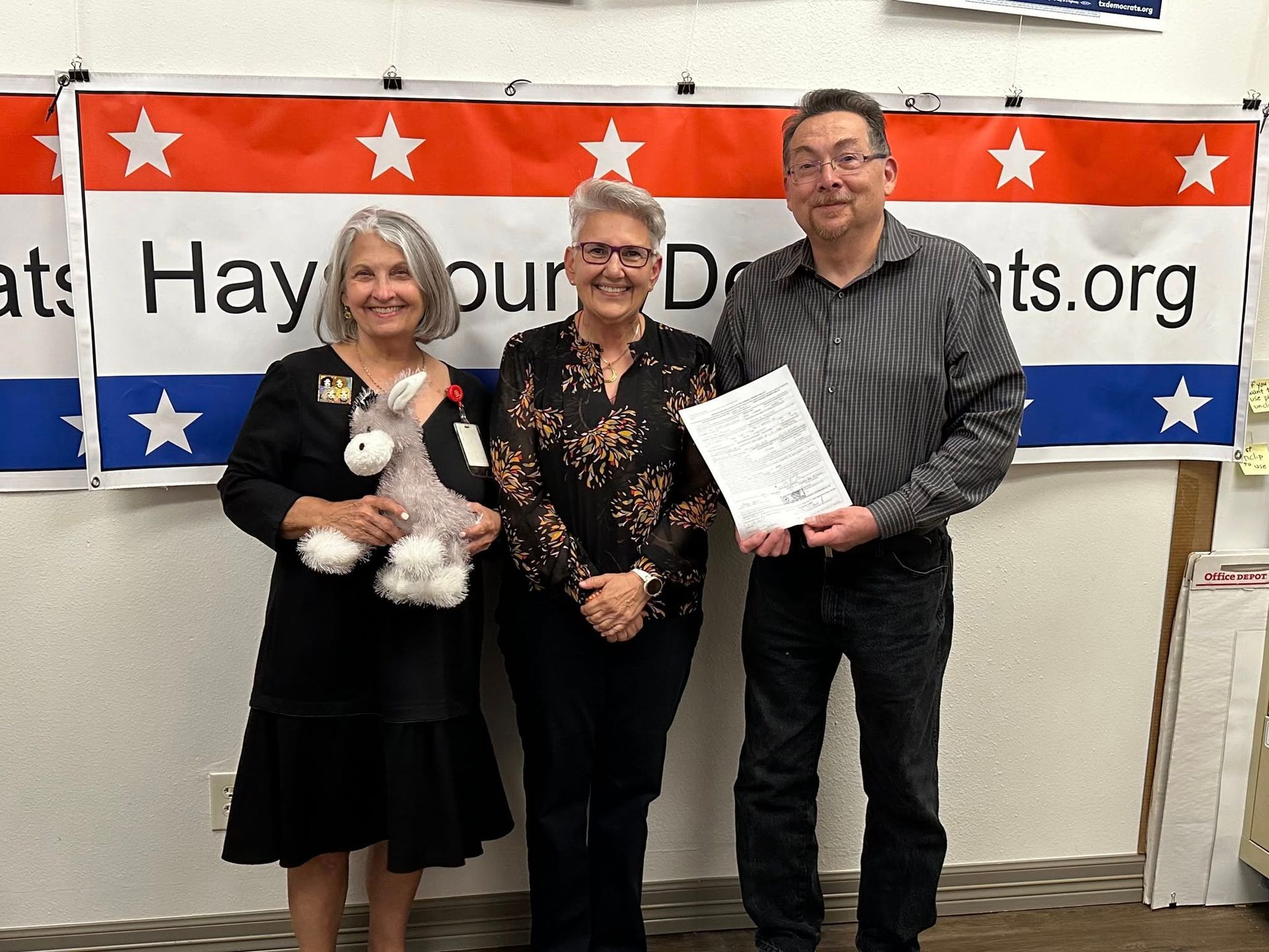 Three people standing in front of a banner; two women and a man. The man holds a paper.