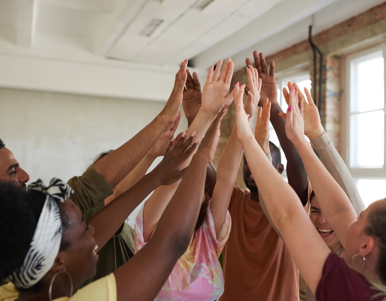 A diverse group of people stand in a circle with their arms raised, hands meeting in the center to celebrate.
